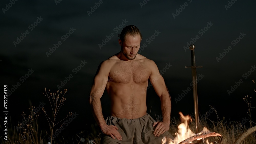 Shirtless male warrior sitting on knees near burning fire with sword ...