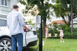 Modern and environmental conscious man recharge electric vehicle from home charging station on while his playful family playing together in background. Energy sustainable for better future. Synchronos