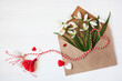 Envelope with spring flowers snowdrops and red and white martenitsa, tassel cord, a symbol of the Martisor holiday, Baba Marta on a white wooden background. Greeting card.