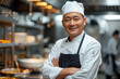 © Enrique - A middle-aged Chinese chef wearing a chef's hat and arms crossed is wearing an apron standing in the restaurant kitchen and smiling, his eyes against the camera