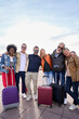 © CarlosBarquero - Vertical portrait group mature tourist friends embraced in affectionate attitude looking smiling camera together on vacation. Diverse joyful tourism people with luggage in city street. Top copy space