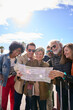 © CarlosBarquero - Vertical group diverse middle-aged tourist people standing looking at travel map in hands on street vacation city smiling on sunny day. Adults community friends enjoying trip together. Top copy space
