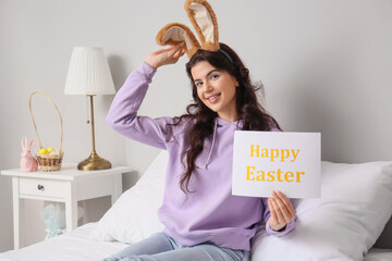 Canvas Print - Young woman in bunny ears holding card with text HAPPY EASTER at home