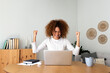 © Daniel - Happy young multiracial Hispanic female celebrating success with arms raised up in front of laptop at home office.
