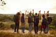 © Studio Romantic - Back view of a group of men and women hikers with large backpacks looking into the distance waving hands during hiking in nature standing in a row together. Friends on a trekking trip holidays.