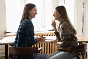 Wall Mural - Happy young female business friends talking at workplace table, having fun, laughing, discussing funny creative ideas for project, enjoying office friendship, teamwork, cooperation