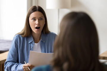 Shocked entrepreneur woman holding paper document, staring at female business colleague, project partner with open mouth. Job candidate surprising employer at interview