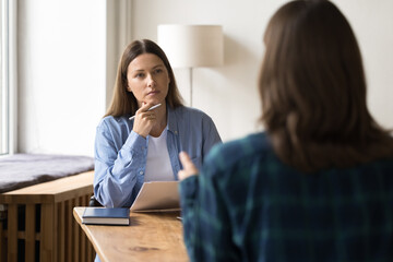 Wall Mural - Serious female employer listening to candidate at job interview, sitting at work table, touching chin, holding paper resume. HR recruit manager woman meeting with applicant