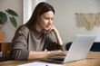 © fizkes - Positive pensive freelance professional woman typing on laptop in home office , sitting at table, touching chin, looking at display, working on business project, enjoying online communication