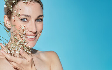 Wall Mural - Skin care concept. Beauty portrait of smiling Caucasian woman with flowers near healthy moisturized facial skin, isolated on blue background