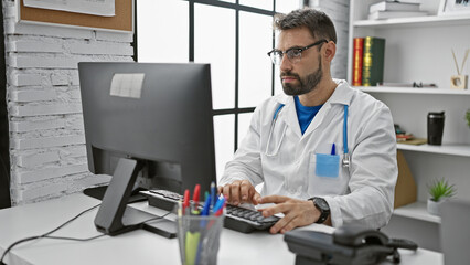 Bearded young hispanic man doctor hard at work, using his computer indoors at a medical clinic