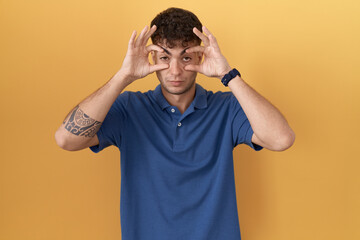 Sticker - Young hispanic man standing over yellow background trying to open eyes with fingers, sleepy and tired for morning fatigue