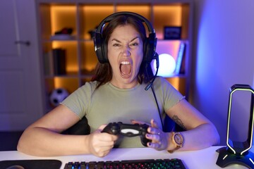 Poster - Beautiful brunette woman playing video games wearing headphones angry and mad screaming frustrated and furious, shouting with anger. rage and aggressive concept.