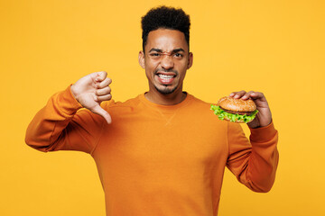Wall Mural - Young displeased man wear orange sweatshirt casual clothes eat burger show thumb down gesture isolated on plain yellow background studio. Proper nutrition healthy fast food unhealthy choice concept.