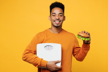 Wall Mural - Young man of African American ethnicity wear orange sweatshirt casual clothes eat burger hold scales isolated on plain yellow background. Proper nutrition healthy fast food unhealthy choice concept.