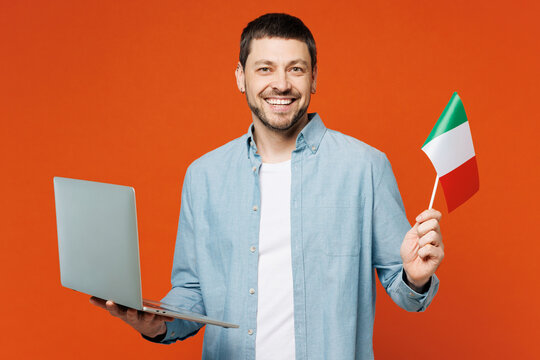 Young IT man he wears blue shirt white t-shirt casual clothes hold Italian flag use work on laptop pc computer look camera isolated on plain red orange background studio portrait. Lifestyle concept.