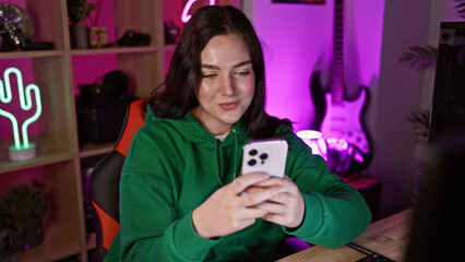 Wall Mural - A young caucasian woman uses a smartphone in a colorful gaming room at night, showcasing technology and leisure activities.