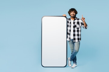 Full body young Indian man wears shirt white t-shirt casual clothes big huge blank screen area mobile cell phone smartphone point finger up isolated on plain blue cyan background. Lifestyle concept.