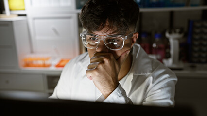 Wall Mural - Handsome, serious-faced young hispanic male scientist engrossed in high-stakes research, working diligently with computer in dim laboratory, amidst glowing test tubes and microscope.