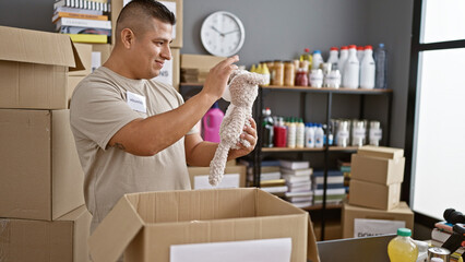 Canvas Print - Joyful latin guy volunteering with a smile, packing teddy bear donations in cardboard at community charity center