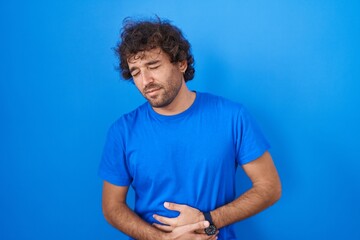 Poster - Hispanic young man standing over blue background with hand on stomach because indigestion, painful illness feeling unwell. ache concept.