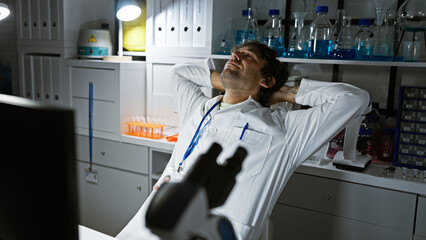 Wall Mural - A tired young man in a white lab coat is taking a break in a chemistry laboratory, surrounded by scientific equipment.