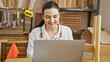 © Krakenimages.com - Smiling young hispanic woman using laptop in a woodworking workshop studio.