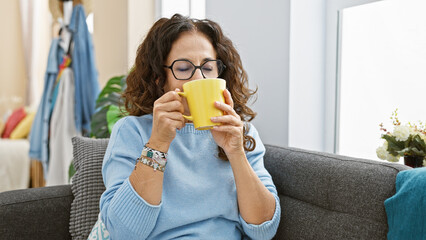 Sticker - Mature hispanic woman enjoys coffee indoors, sitting on a sofa with glasses and curly hair.