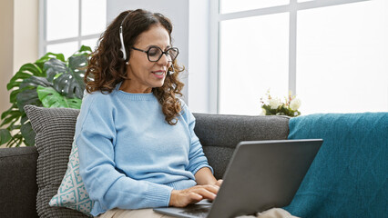 Wall Mural - A smiling middle-aged hispanic woman with curly hair working on a laptop in a bright living room at home, radiating a sense of comfort and productivity.