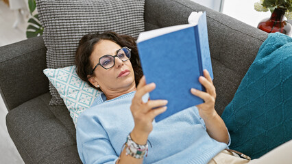 Sticker - Mature hispanic woman reading a book, relaxing on a sofa indoors, surrounded by home decor.