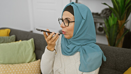 Canvas Print - A mature woman wearing glasses and a hijab speaks into a smartphone in a cozy living room.