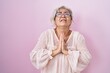 Middle age woman with grey hair standing over pink background begging and praying with hands together with hope expression on face very emotional and worried. begging.