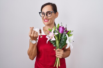 Sticker - Middle age brunette woman wearing apron working at florist shop holding bouquet doing money gesture with hands, asking for salary payment, millionaire business