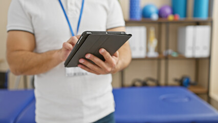 Canvas Print - Young hispanic man with a beard, wearing lanyard, using tablet in a rehab clinic interior.