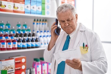 Canvas Print - Middle age grey-haired man pharmacist talking on smartphone reading prescription at pharmacy