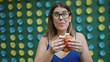 Beautiful hispanic woman in glasses delightfully eating traditional japanese melon pan snack on tokyo's vibrant street