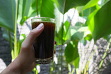 hand holding a glass of coffee with green plants in the background
