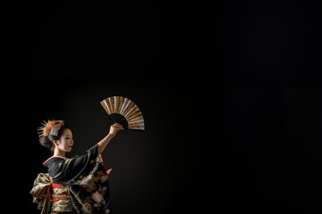 Magnifique Geisha vêtue d'un kimono en soie traditionnel et d'une coiffure ancestrale avec des peignes à chignon exécutant la danse de l'éventail sur fond noir avec espace négatif pour texte copyspace