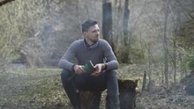 Lonely Thoughtful Tourist Sits On Stump In The Forest With Ax And Looks Around. Hispanic Middle Aged Man Is Resting In Wild. Smoke From Fire Spreads Over Ground In Front Of The Person.