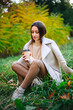© Vadim - Young beautiful girl sits on the grass in white coat and sweater and drinks coffee in the autumn park.