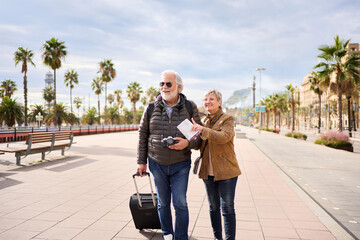 Wall Mural - Older Caucasian tourist couple smiling looking and pointing interest places. Elderly woman and man standing in European city street enjoying pensioner vacation. Senior tourism people traveling happy