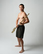 Full length portrait of fit handsome shirtless asian male model,  Holding golden trident weapon, standing in warrior training action pose, isolated on white studio background.