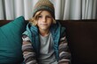 © Iigo - Portrait of a little boy in a warm hat and a plaid sitting on a sofa at home.