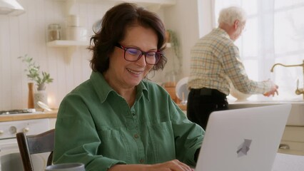 elderly woman sits at table in kitchen with laptop. types on keyboard looks intently in screen, smiles. succeed in using computer and internet, feels pleased and comfortable. old man on background