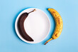 Spoiled yellow and brown bananas on a white plate on a light blue background from above. Food waste and excessive food consumption in the world.