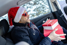 Fashionable Man With Santa Hat And Red Sunglasses Holding Christmas Presents While Sitting On Rear Car Seat. Winter Scene In The Background.