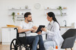 © sofiko14 - Portrait of brunette caucasian female doctor checking up condition of bearded male patient at home. Nurse talking to mature man on wheelchair, showing examination results using modern tablet.
