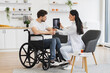 © sofiko14 - Full length portrait of Caucasian woman doctor checking up condition of mature bearded male patient at home. Nurse talking to mature man wheelchair user, showing x-ray scan of spine on modern tablet.