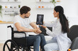 © sofiko14 - Young female nurse talking to mature man wheelchair user, showing x-ray scan of spine on modern tablet. Adult Caucasian woman doctor checking up condition of mature bearded male patient at home.
