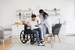 Focused female in doctor's coat supporting bearded male rising from wheelchair indoors. Health professional consulting man after back injury about physio therapy at home.